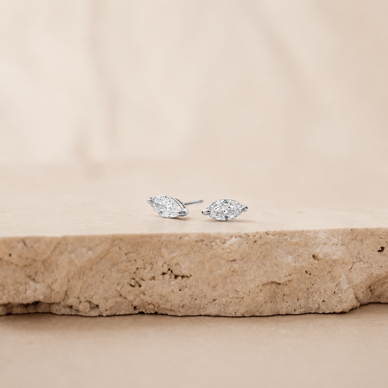 A pair of silver stud earrings with marquise-cut clear gemstones, resting on a textured beige stone slab against a soft beige background.